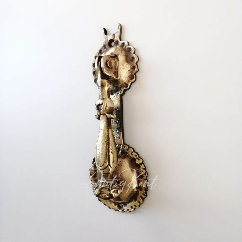 May include: A gold-toned metal door knocker with an ornate design. The knocker has a ring-shaped handle and a decorative base.