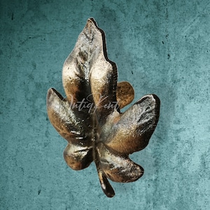 Puede incluir: Un tirador de cajón de metal de color dorado en forma de hoja. La hoja tiene un diseño detallado y una superficie ligeramente texturizada.