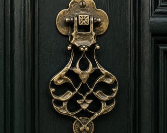 May include: An antique brass door knocker with a weathered finish and intricate scrollwork design. The knocker features a central hinged element attached to a clover-shaped backplate, mounted securely onto a dark green paneled wooden door.