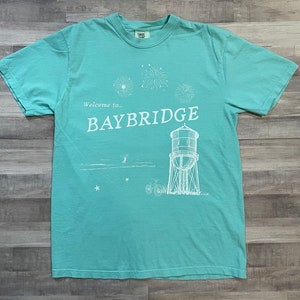 May include: A teal blue t-shirt with a white graphic design of a water tower, fireworks, and the text "Welcome to... BAYBRIDGE".