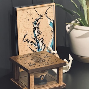 May include: A wooden shadow box with a navy blue hat inside. The box has an engraved inscription that reads "C.T.C. ROBERT T. GODWIN FY 22 CLASS 130". The box is sitting on a table next to a wooden map of the Chesapeake Bay.