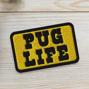 May include: A yellow and black embroidered patch with the words "PUG LIFE" in bold, black letters. Two small potted succulents are placed near the patch on a light wood-grain surface. The patch has a black border.