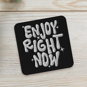 May include: Black square patch with white embroidered text that reads "ENJOY RIGHT NOW" on a white wooden surface with two green succulent plants in white pots.