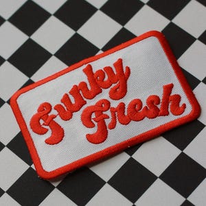 Könnte beinhalten: Ein rechteckiger Aufnäher mit den Worten "Funky Fresh" in roter Schrift auf weißem Grund. Der Aufnäher hat einen roten Rand und ist auf einem schwarz-weißen Schachbrettmuster angeordnet.