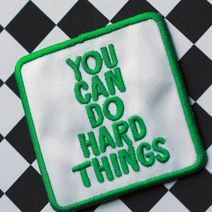 May include: A square embroidered patch with a white background and a green border. The patch features the green embroidered text "YOU CAN DO HARD THINGS" in a stacked, sans-serif font, centered within the square frame with rounded corners.