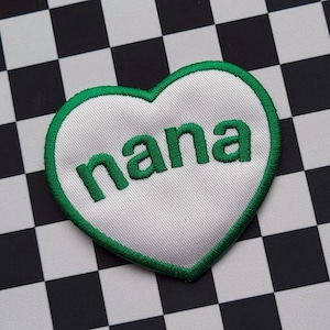 Könnte beinhalten: Ein weißer herzförmiger Patch mit grünen Nähten, mit dem Wort "nana" in grüner Stickerei auf einem schwarz-weiß karierten Hintergrund.