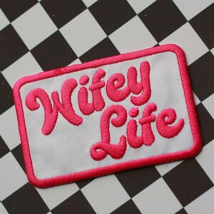 May include: A rectangular embroidered patch with a white background and a bright pink border. The text Wifey Life is stitched in a bold, cursive, bright pink font across the center of the patch, creating a clear and readable design.