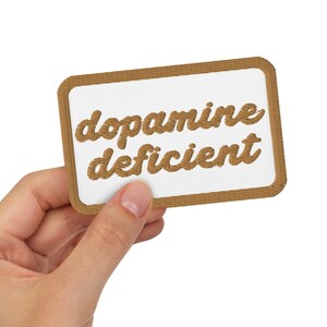 May include: A hand holds up a brown rectangular patch with white text that reads "dopamine deficient" in a cursive font.