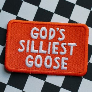 Pode incluir: Um patch retangular laranja com o texto bordado "GOD'S SILLIEST GOOSE" em branco. O patch está sobre um fundo xadrez preto e branco.