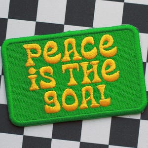 May include: Green embroidered patch with the words "peace is the goal" in yellow, stylized lettering. The patch has a green border and is set against a black and white checkered background. Ideal for jackets, bags, or other fabric items.