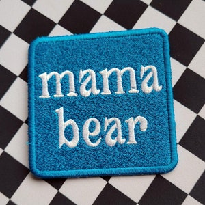 May include: A square, turquoise patch with the words "mama bear" embroidered in white. The patch is set against a black and white checkered background.