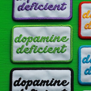 May include: Three white fabric patches with black, green, and purple stitching. Each patch reads "dopamine deficient" in a cursive font.
