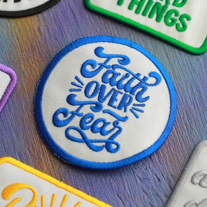 May include: Embroidered patches with motivational phrases. A circular patch with the words "Faith Over Fear" in blue script is framed by a blue border. Other patches include the words "Good Things" and "Dopamine".