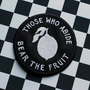 May include: A circular black embroidered patch featuring a white stitched lemon graphic in the center. The text "THOSE WHO ABIDE BEAR THE FRUIT" is embroidered in white capital letters around the lemon, following the circular edge of the patch.