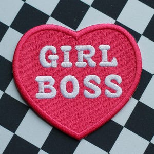 May include: A pink heart-shaped embroidered patch with the words "GIRL BOSS" in white lettering. The patch is set against a black and white checkered background, creating a bold and eye-catching design.