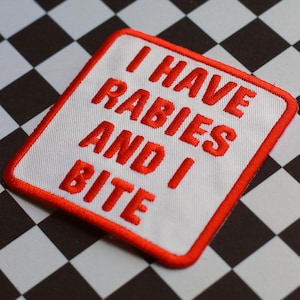 Puede incluir: Un parche bordado cuadrado con fondo blanco y un borde rojo grueso. El parche presenta el texto I HAVE RABIES AND I BITE bordado en letras mayúsculas rojas y negritas en el centro, creando un diseño claro y de alto contraste.