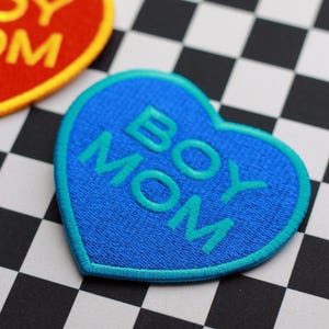 May include: A blue heart-shaped patch with the words "BOY MOM" embroidered in turquoise. The patch has a turquoise border and is set against a black and white checkered background. A red heart-shaped patch with yellow text is partially visible.