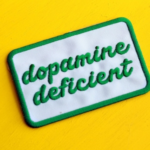 Puede incluir: Un parche blanco con un borde verde y el texto "dopamine deficient" bordado en verde.
