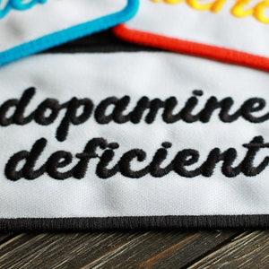 Pode incluir: Um emblema retangular branco bordado com uma borda preta. O texto 'dopamine deficient' está bordado em letra cursiva preta no centro. Dois outros emblemas parciais com texto azul e amarelo são visíveis na parte superior.