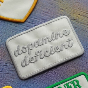 Può includere: Una toppa rettangolare ricamata con sfondo bianco e bordo grigio. Il testo "dopamine deficient" è cucito in corsivo grigio al centro della toppa. La toppa presenta angoli arrotondati e una texture in tessuto pulita e ben definita.