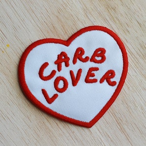 Pode incluir: Um remendo branco em forma de coração com contorno vermelho e as palavras "CARB LOVER" bordadas em vermelho.