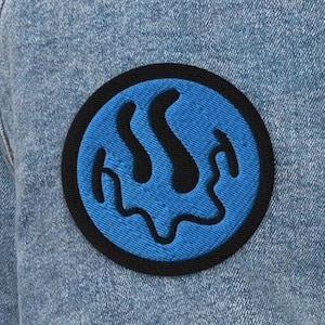 May include: A blue denim jacket with a black and blue embroidered patch on the left sleeve. The patch features a blue circle with a black dripping design inside.