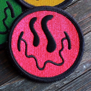 Puede incluir: Tres parches bordados en forma de cara sonriente con ojos que gotean. Los parches son amarillos, verdes y rosas.