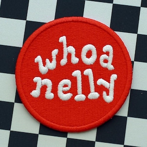 Puede incluir: Un parche redondo rojo con texto bordado en blanco que dice "whoa nelly" sobre un fondo a cuadros blanco y negro.