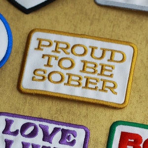 May include: A collection of embroidered iron-on patches with various religious and inspirational messages. The patches are in different colors and shapes, including circles, squares, and hearts. The text on the patches includes phrases like "Faith Over Fear", "Love Like Jesus Does", "Proud to Be Sober", and "Praise the Lord".