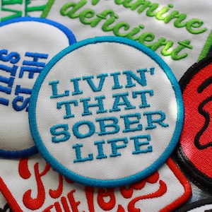 May include: A collection of embroidered patches with various sayings. One patch is white with blue text that reads "Livin' That Sober Life".