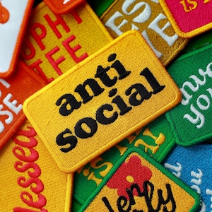 Op de afbeelding: Een verzameling geborduurde patches in verschillende kleuren, waaronder geel, oranje, groen en roze. De patches tonen zinnen als "anti social", "love like Jesus" en "garden girly". De patches hebben een getextureerde uitstraling.