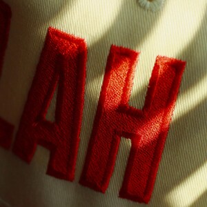 Peut inclure: Gros plan sur une casquette de baseball beige avec les lettres rouges brod&eacute;es "LAH". La casquette est faite d'un tissu textur&eacute; et est partiellement &eacute;clair&eacute;e par la lumi&egrave;re du soleil, cr&eacute;ant des ombres.