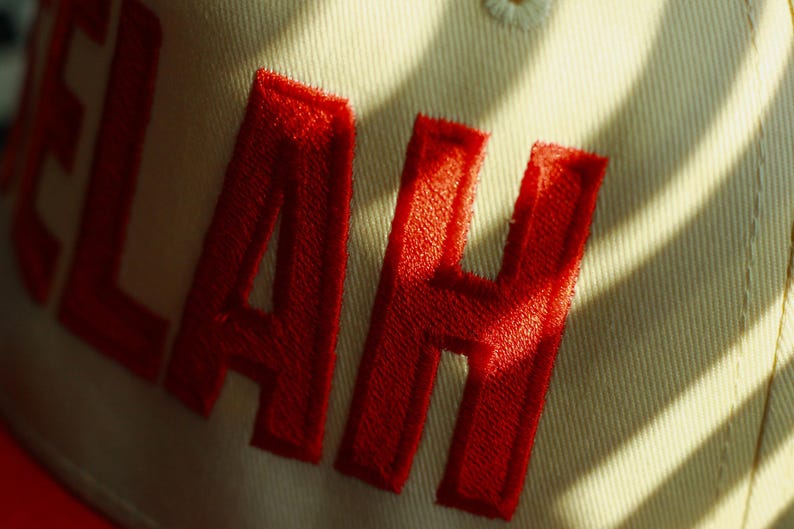 Peut inclure: Gros plan sur une casquette de baseball beige avec les lettres rouges brod&eacute;es "LAH". La casquette est faite d'un tissu textur&eacute; et est partiellement &eacute;clair&eacute;e par la lumi&egrave;re du soleil, cr&eacute;ant des ombres.