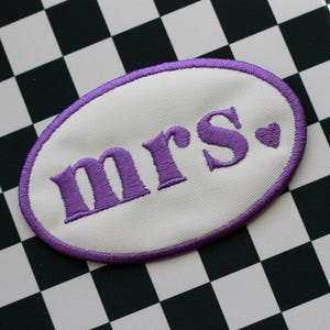Op de afbeelding: Een witte ovale patch met paarse borduurwerk met de tekst "mrs." met een klein paars hartje.