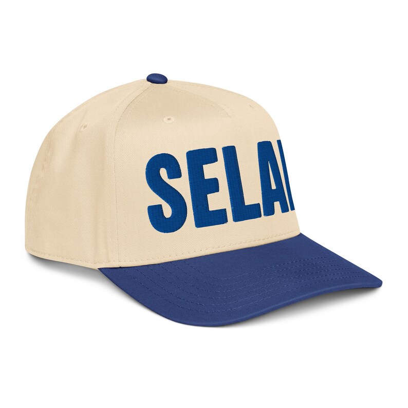 Peut inclure: Casquette de baseball avec une couronne cr&egrave;me et une visi&egrave;re bleu marine. Le mot "SELA" est brod&eacute; en lettres bleu marine sur le devant. La casquette a une visi&egrave;re incurv&eacute;e et un bouton sur le dessus.
