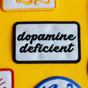May include: White patch with black embroidered text that reads "dopamine deficient" with a black border. The patch is on a yellow background.