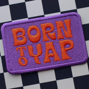 Könnte beinhalten: Ein rechteckiger, lila bestickter Aufnäher mit den Worten "BORN TO YAP" in orangefarbenem Faden. Der Aufnäher ist vor einem schwarz-weiß karierten Hintergrund platziert.