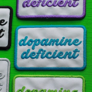 May include: Four white fabric patches with blue, purple, black, and green stitching. The patches have the text "dopamine deficient" in blue, "serotonin deficient" in purple, "serotonin deficient" in black, and "dopamine deficient" in green.