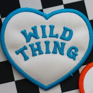 Op de afbeelding: Witte hartvormige patch met een blauwe rand en de tekst "WILD THING" geborduurd in blauw.