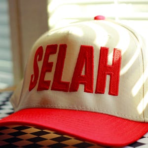 Peut inclure: Casquette de baseball beige avec une visi&egrave;re rouge. Le mot "SELAH" est brod&eacute; en lettres rouges sur le devant. La casquette est pos&eacute;e sur une surface &agrave; carreaux noirs et blancs.