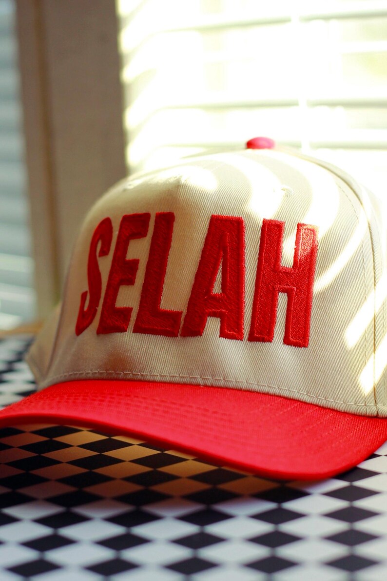 Peut inclure: Casquette de baseball beige avec une visi&egrave;re rouge. Le mot "SELAH" est brod&eacute; en lettres rouges sur le devant. La casquette est pos&eacute;e sur une surface &agrave; carreaux noirs et blancs.