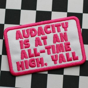 May include: Embroidered patch with the phrase "AUDACITY IS AT AN ALL-TIME HIGH, Y'ALL" in bright pink lettering on a white background. The patch has a pink border and is set against a black and white checkered pattern.