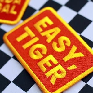 May include: Embroidered patch with the words "EASY TIGER" in yellow on a red rectangular background with a yellow border. The patch is set against a black and white checkered background. Another patch is visible in the background.