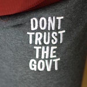 Può includere: Primo piano di pantaloni della tuta grigio scuro con la scritta ricamata "DONT TRUST THE GOVT" in bianco. La parte superiore di una maglietta rossa a coste è visibile sopra i pantaloni. L'immagine è ben illuminata.