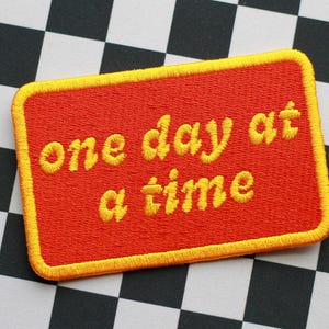 May include: Embroidered patch with the phrase "one day at a time" in yellow script on a red rectangular background. The patch has a yellow border and is set against a black and white checkered pattern.