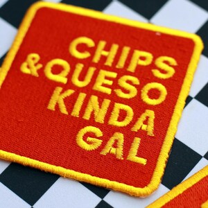 Può includere: Una toppa quadrata rossa con testo ricamato giallo che recita "CHIPS & QUESO KINDA GAL". La toppa ha un bordo giallo ed è posta su uno sfondo a scacchi bianco e nero.