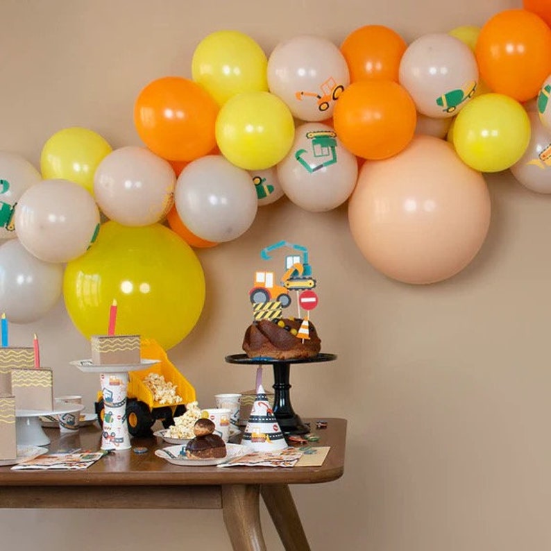 May include: A construction-themed birthday party with a yellow and orange balloon arch, a cake with construction vehicle toppers, and a table set with construction-themed party supplies.