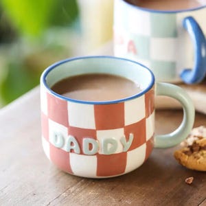 Op de afbeelding: Een keramische mok met een rood-wit geblokt patroon, gevuld met een bruine vloeistof. Het woord "DADDY" staat in lichtblauwe letters op de voorkant. De mok heeft een lichtgroen oor en een blauwe rand. Een tweede mok is zichtbaar op de achtergrond.