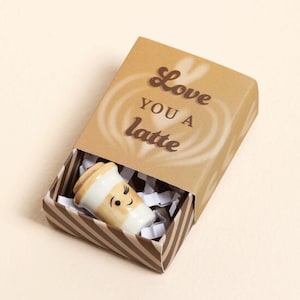 May include: A miniature ceramic coffee cup with a smiling face, presented in a brown and white striped box. The box lid features the words "Love You a Latte" in brown. The box is filled with white paper for cushioning.