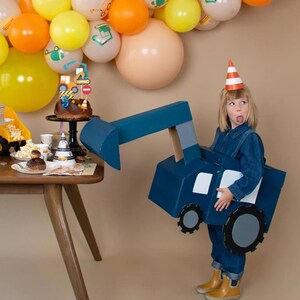 May include: A child wearing a blue construction vehicle costume, holding a cardboard digger, with a birthday cake and decorations in the background.